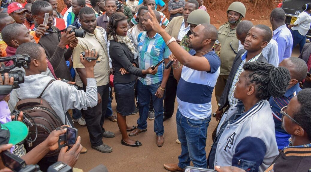 Crowd surrounds journalists recording a heated public discussion outdoors